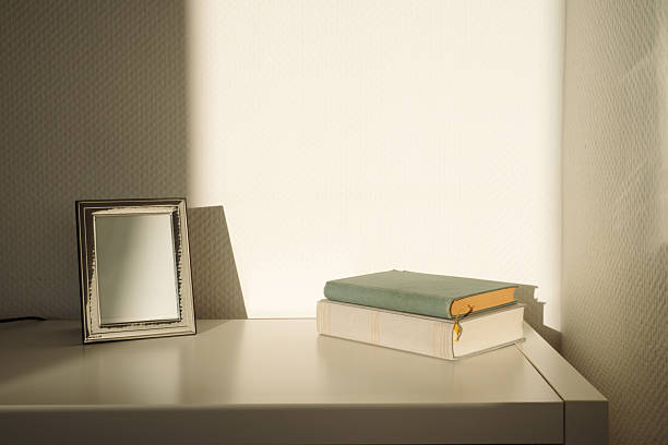 istock Empty silver photo frame and old books in direct sunlight on a white table 2251511815