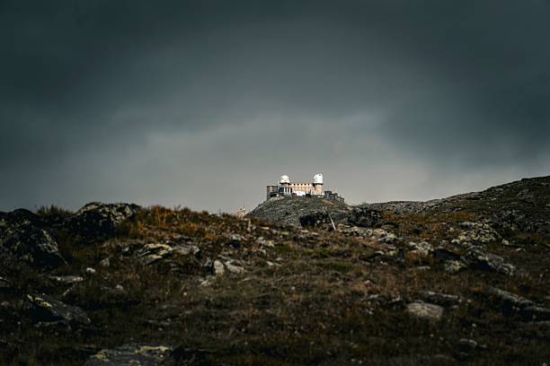 A remote astronomical observatory, featuring prominent white domes, stands atop a rugged, rocky mountain peak under a dramatic