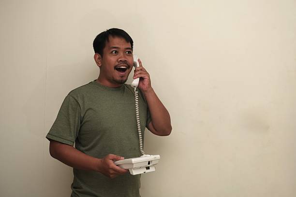 Portrait of a Southeast Asian man with a shocked expression while using a landline phone, Isolated, perfect for communication or ad use.