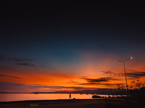 Scenic view of a solitary figure walking along the waterfront promenade at dusk. The sky features a vibrant gradient of orange and deep blue colors over the calm sea and distant mountains, illuminated by a street lamp.