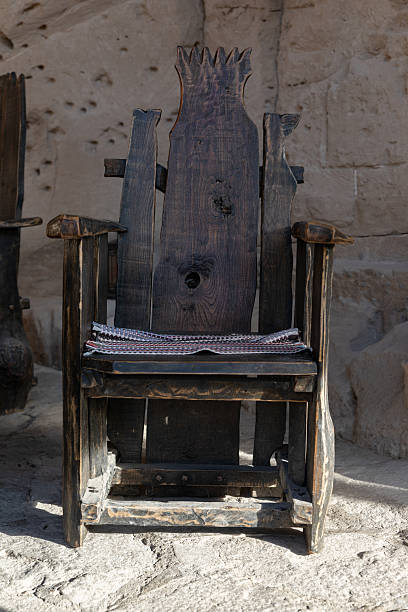Handcrafted dark wood chair with a distinct backrest design, placed against a stone wall in natural sunlight, highlighting its texture and craftsmanship.
