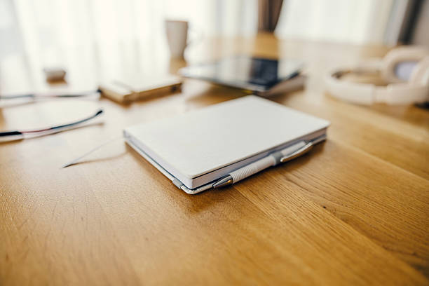 Workspace desk with a closed white notebook and pen, complemented by blurred headphones, eyeglasses, and a smartphone, highlighting a calm home office environment for work or study