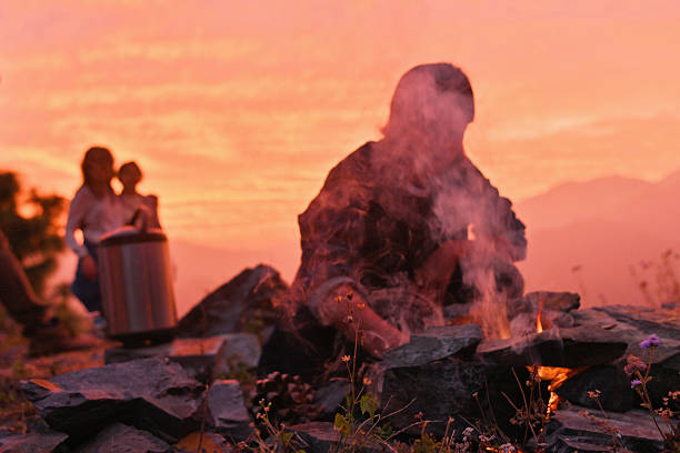 A family camping scene set in the mountains at sunset, featuring a person tending a campfire built with stones while smoke rises into the warm evening sky. Other family members stand in the background near cooking equipment, creating a cozy and authentic outdoor atmosphere. The image represents family bonding, camping lifestyle, outdoor cooking, adventure travel, and connection with nature.