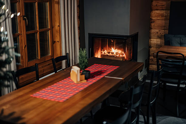 Cozy, luxurious, and empty modern living room with two beige textile sofas, three low stools on the hairy carpet, and decoration (pillows, blankets, books, eyeglasses) on the hardwood floor in front of a design-shaped modern fireplace on an anthracite black plaster wall and copy space in the middle. A long marble shelf under the fireplace with decorations, logs, candles, and a vase of dry flowers. Large and high windows with blinds and with a terrace and garden view (chairs, tables, and trees) on both sides of the fireplace. A slight vintage effect was added. 3D rendered image.