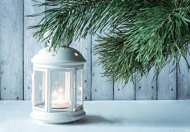 white vintage lantern with burning candle and green pine branches on a white wooden background