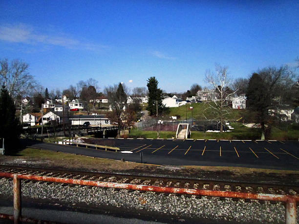 Quiet railroad-side scene in Bridgeport, West Virginia, where gravel-lined tracks meet a suburban parking lot. Leafless trees and clear skies suggest early spring or late fall. Residential homes and parked vehicles frame the moment with everyday rhythm and small-town stillness. Bridgeport WV, USA February 07, 2016