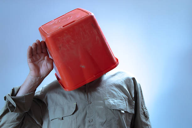 A man with a red plastic bucket on his head puts his hand on his ear to hear more clearly
