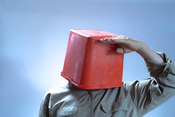 A man with a red plastic bucket on his head puts his hand on his forehead pretending to see further