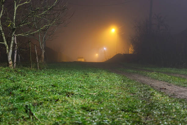 ​A low-angle nighttime photograph showing a rural road leading into thick fog, dramatically illuminated by an orange street lamp.