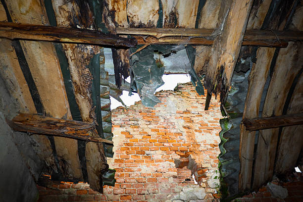 istock ​A view from below shows a ruined roof where a section of an old brick wall is visible through rotted wooden beams and damaged slate. 2251550104