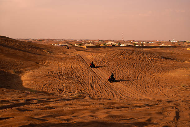 Two riders descend a steep, heavily tracked sand dune on quad bikes, with warm sunset tones, vast desert scenery, and a distant safari camp on the horizon.