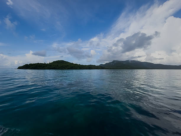 istock Calm ocean seascape with distant green island and mountain range under dramatic cloudscape, blue water surface and wide horizon 2251552027