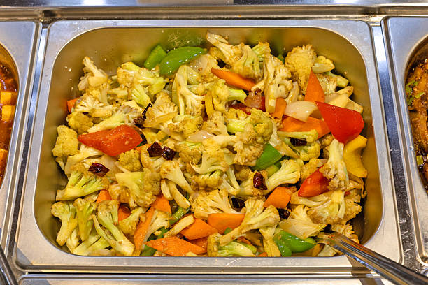 A close-up view of a steaming metal pan filled with a colorful mix of stir-fried cauliflower florets, carrots, bell peppers, and onions, ready to be served