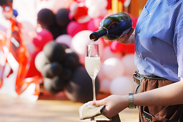 A person in a striped blue shirt pours sparkling drink into a flute glass during a party decorated with colorful balloons.