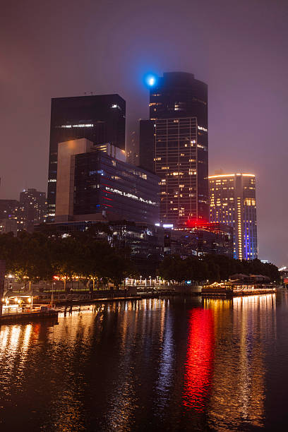A nighttime cityscape along the river shows tall modern buildings glowing with neon lights; Reflections ripple on the water, creating a moody, atmospheric scene perfect for urban, nightlife