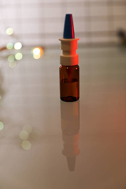 Close-up image of a small amber nasal spray bottle lying on a reflective surface, illuminated by soft red and green lights with a blurred bokeh background.