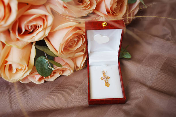 A delicate gold cross pendant rests in an open red jewelry box, surrounded by soft peach roses against a textured fabric background, suggesting a special occasion.