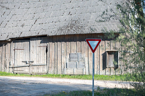 The paint on the structures is weathered, and many windows and doors appear worn. Overgrown plants and weeds line the path and edges of the buildings, suggesting the area is not in active use. A metal overhead structure with cables connects the buildings in the background.