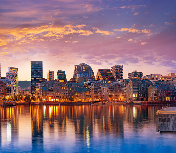 Modern waterfront apartments in Oslo Bispevika at summer sunset with warm lights reflecting on calm harbor water.