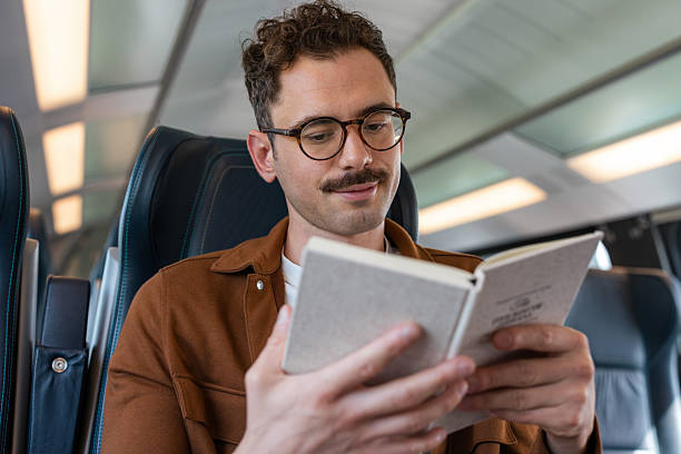 istock Young man with glasses reading a book while traveling on a train 2251587785