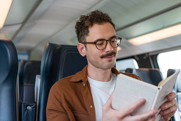 istock Young man with glasses reading a book while sitting on a train 2251587850