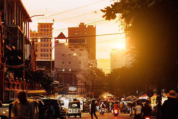 Golden sunset bathes a busy Phnom Penh street, with scooters, cars, and pedestrians framed by urban buildings and overhead cables. A warm, bustling city vibe captures daily life in Cambodia.