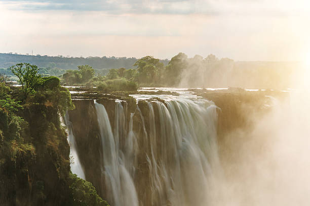 An African 25-29 year old Woman with a black backpack on her back standing looking at camera with Victoria Falls Mosi-oa-Tunya at sunrise Zimbabwe Africa