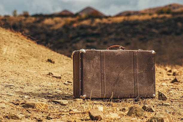 A brown suitcase is sitting on a rocky, dry ground. The scene is desolate and barren, with no signs of life or vegetation. The suitcase is the only object in the image