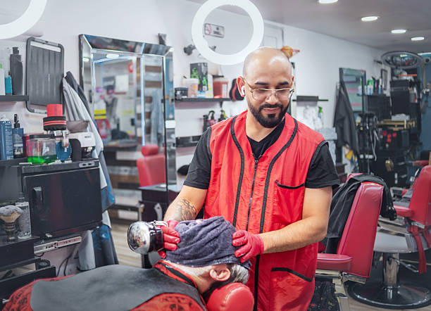 Bald barber with a beard and glasses uses a handheld massager on a client's head in a barbershop. The client is seated in a red barber chair with a dark towel over his head. The barber, wearing a red vest and pink gloves, is focused on the task. The background shows a fully equipped salon with shelves of products, mirrors, and ring lights. Medium shot with an eye level view.