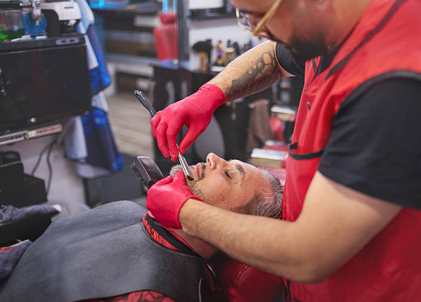 Barber with a tattooed arm wears pink gloves while carefully shaving a mature man's face with a straight razor. The client is leaning back in a barber chair with a protective cape on, his eyes are closed during the grooming service. The scene takes place in a barbershop during the afternoon. High angle view with selective focus on the shaving action.