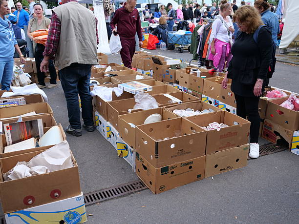 Flea market scene with kitchenware, toys, books, and electronics displayed on pavement. Vendors selling second-hand goods outdoors.