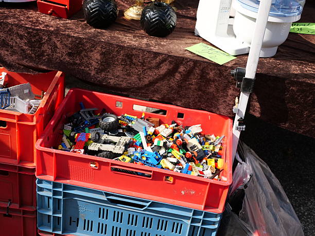 second hand Lego bricks mixed box for sale at flea market stall, assorted toy building blocks in red plastic crate, used vintage toys, large black monster truck tires, outdoor market goods.