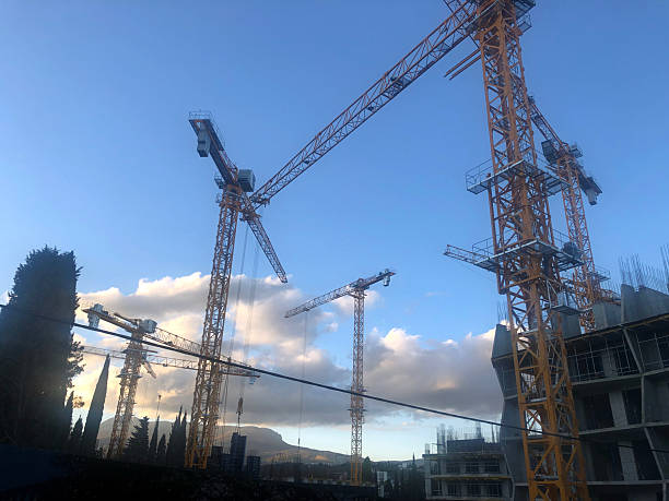 Construction site with multiple yellow cranes against a blue sky. Clouds are visible in the background. The scene represents urban development and infrastructure.