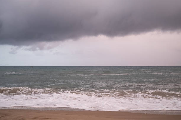Grey storm clouds roll over a calm ocean as gentle waves break on wet sandy shore, creating a moody, tranquil seascape with dramatic overcast sky and distant horizon
