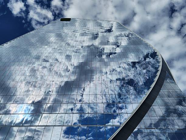 Low angle view of a modern glass skyscraper reflecting clouds and blue sky. Contemporary architecture, urban design and abstract geometric patterns.