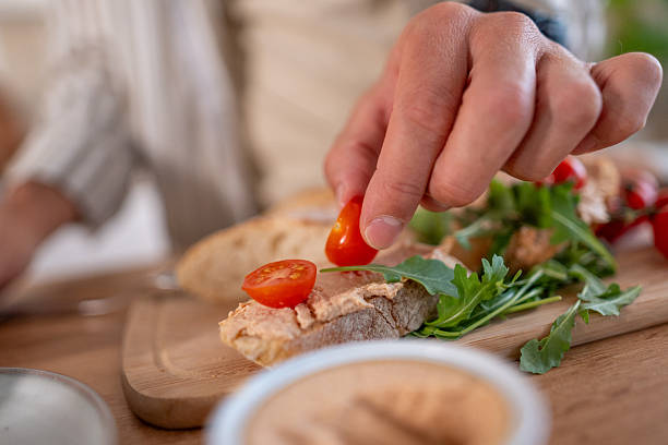 Hand arranging cherry tomato halves on artisanal bread with hummus, arugula and fresh ingredients on a wooden board, creating a healthy homemade bruschetta snack