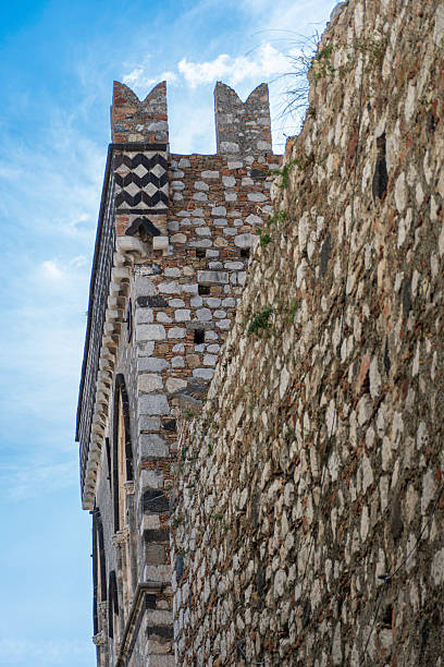 Medieval stone tower and fortified palace façade in Taormina, Sicily, Italy, featuring crenellated walls and Gothic-style windows under a blue sky, showcasing historic architecture and cultural heritage of southern Italy.