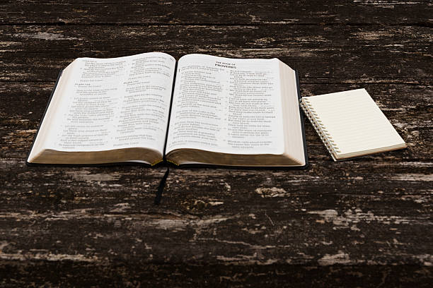 Flat lay of open Holy Bible (Psalms 149-150, Proverbs 1) and notepad on rustic wood table. Top view in natural daylight. Symbolizes spiritual study, wisdom, reflection, and devotion.