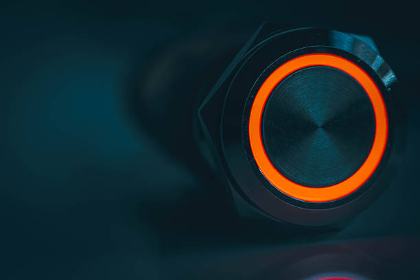 Close-up of a stainless steel push button illuminated with a bright red LED ring, captured against a dark blue background. The cinematic lighting and metallic texture create a futuristic, high-tech feel. Ideal for themes related to electronics, industrial controls, cyber interfaces, technology hardware, and illuminated switches.