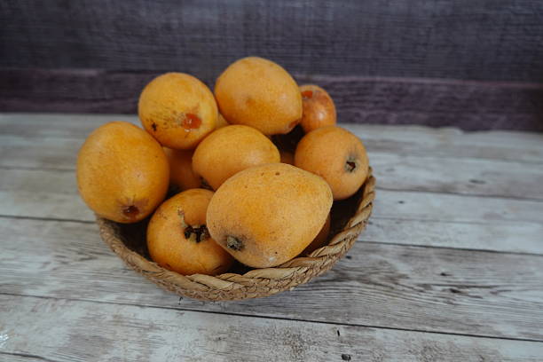 Ripe loquats, also known as japanese medlars or japanese plums, sit in a wicker basket on a weathered wooden surface