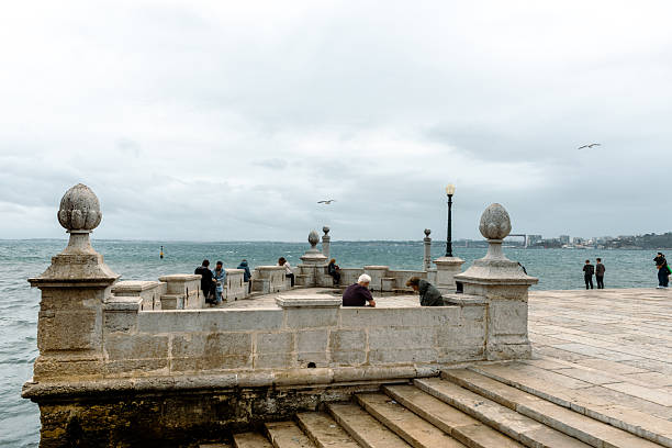 Lisbon. A stone waterfront promenade with scattered visitors gazing at the choppy sea under an overcast sky, capturing a calm yet windswept coastal atmosphere.