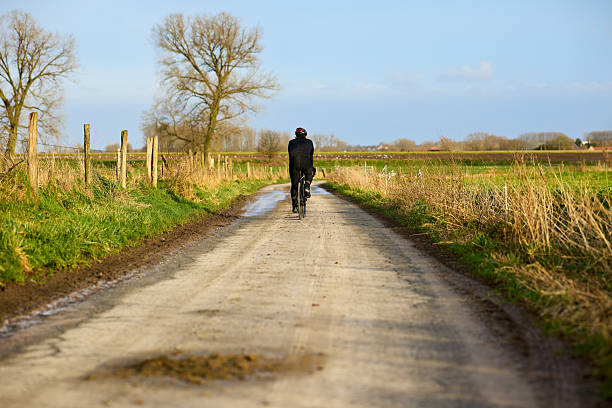 istock A lone cyclist pedals down a muddy rural track, surrounded by golden fields and bare trees under a clear blue sky. A peaceful scene of outdoor recreation in nature. 2251698385