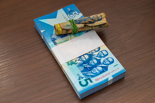istock A crisp, plastic-wrapped stack of new blue 5 Ghana Cedi banknotes sits next to a small, worn, rolled bundle of 5 Cedi notes secured by a green rubber band on a dark wood surface. 2251705459