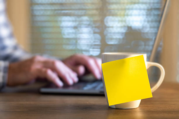 Yellow sticky note on coffee mug with blurred hands typing laptop, home office reminder concept with copy space for message