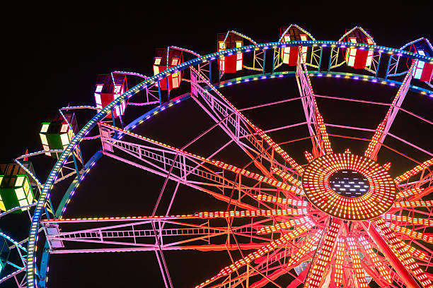 A ferris wheel at night becomes a dazzling spectacle against the dark sky. Its massive circular frame glows with colorful LED lights, often pulsing or changing in vibrant patterns—blues, reds, purples, and greens dancing in rhythmic harmony