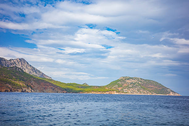 A wide coastal landscape featuring green mountain slopes and rocky cliffs stretching along the shoreline, surrounded by calm deep-blue sea. Above, a dramatic sky with layered white clouds adds depth and atmosphere to the scene. This natural seascape conveys freedom, tranquility, and untouched coastal beauty, making it ideal for travel, nature, outdoor adventure, and lifestyle themes