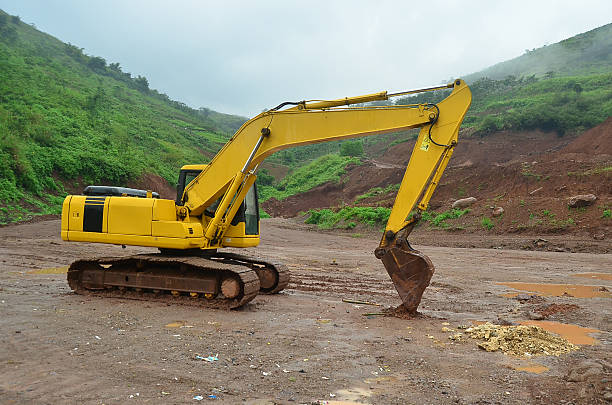 Yellow excavator working at an open construction site surrounded by green hills and soil terrain. Heavy construction machinery used for earthmoving, land clearing, and infrastructure development in outdoor environments.