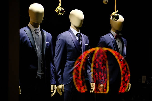 Night view of three headless mannequins dressed in formal men’s suits displayed in a retail store window, with hanging decorative ornaments and colorful light reflections on the glass, set against a dark background with selective lighting.