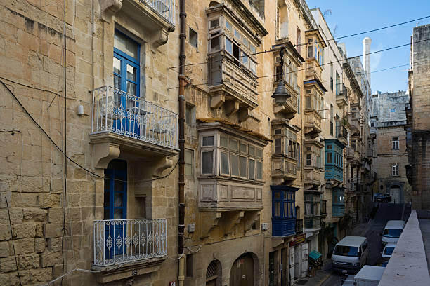 A street in Valletta, Malta, showcases traditional Maltese architecture. The buildings feature enclosed wooden balconies, painted in various colors, adding character to the historic city.