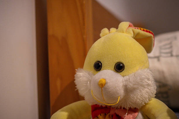 Close-up of a yellow stuffed bunny with button eyes, fluffy cheeks, and a bowtie, resting on a cozy bed indoors.